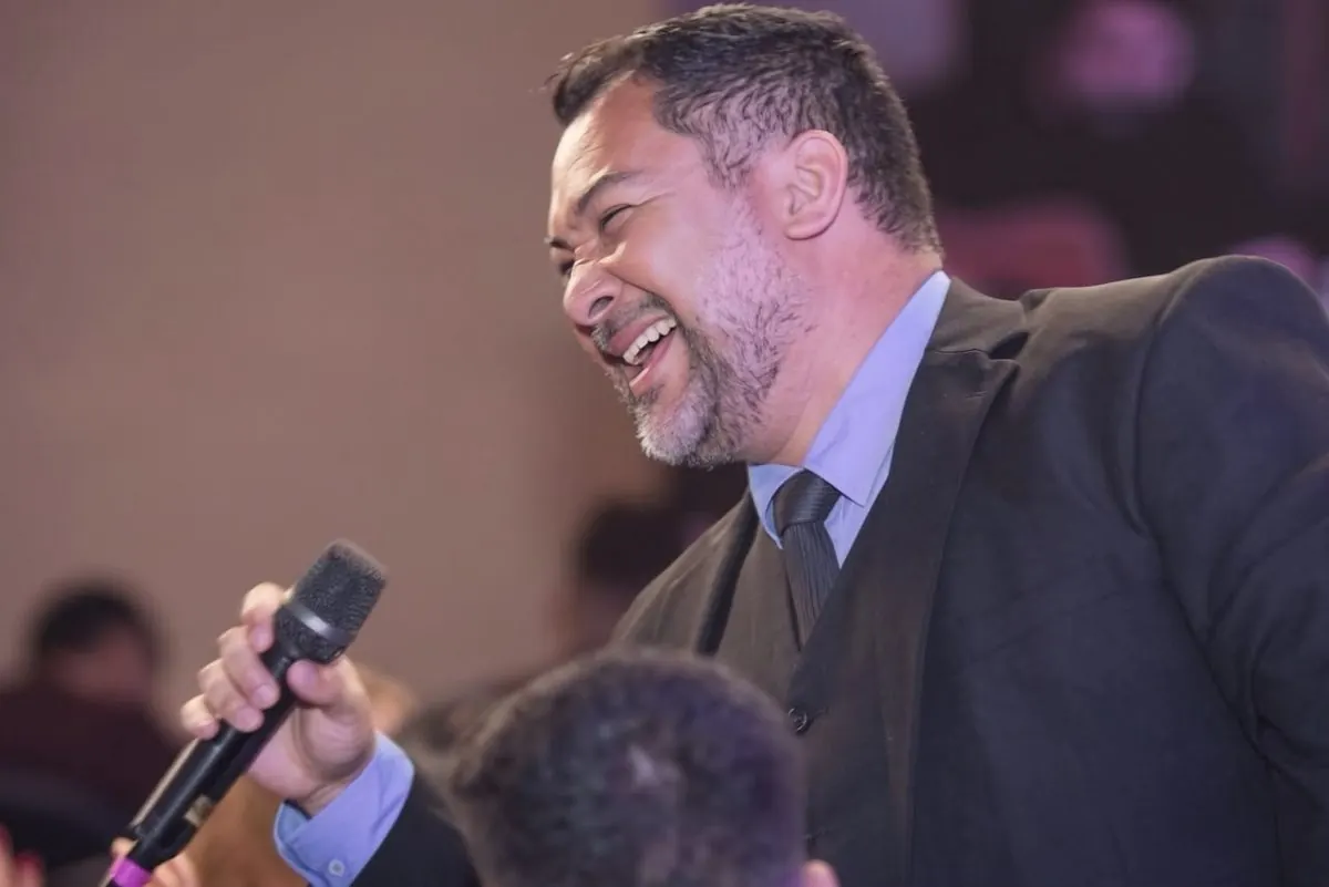 Clayton gargalhando ao microfone, de terno, durante evento.
