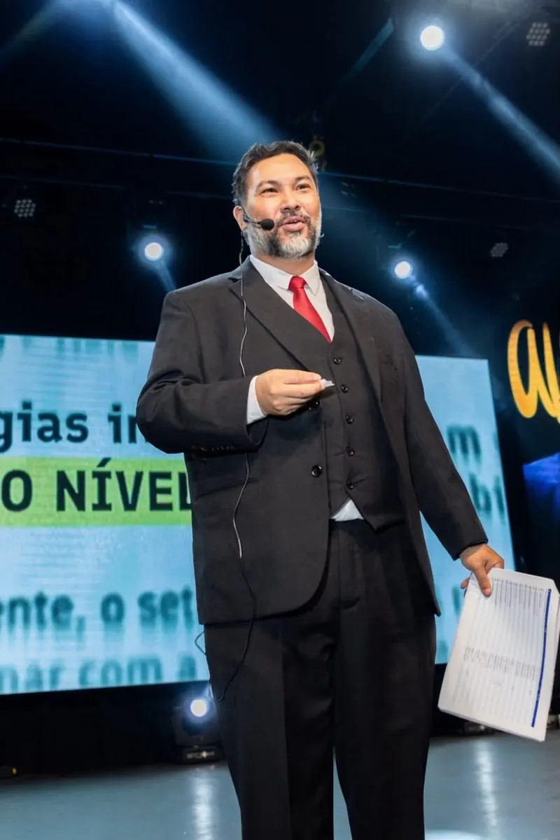 Clayton de terno escuro e gravata vermelha no palco, com microfone de cabeça e roteiro na mão.