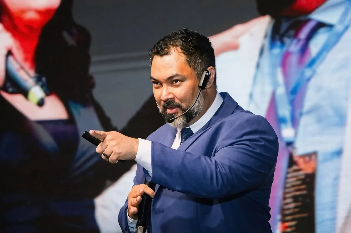 Clayton de terno azul apontando para a plateia, com microfone de cabeça.