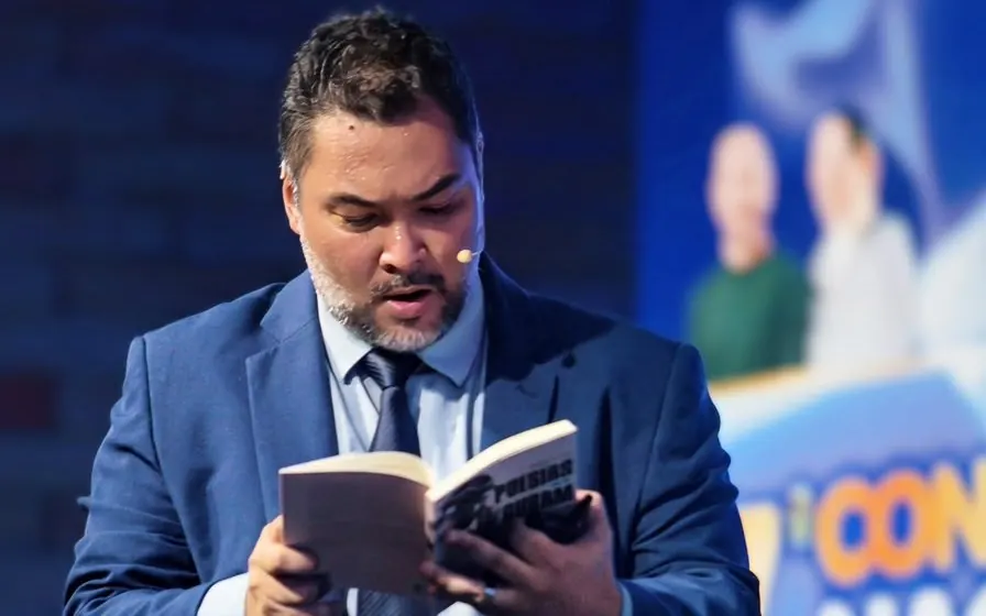 Clayton no palco, de terno azul, lendo um trecho de livro em voz alta para a plateia.