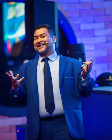 Clayton Yaso no palco, de terno azul e microfone de cabeça, sorrindo com os braços abertos.
