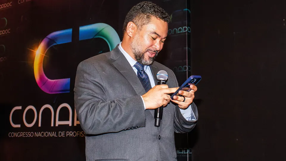 Clayton Yaso no palco do CONAPROC, de microfone na mão, lendo uma mensagem no celular.
