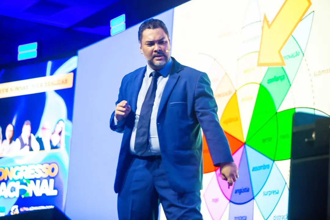 Clayton Yaso no palco do 7º Congresso, de terno azul, apresentando diante de um slide colorido projetado no telão.