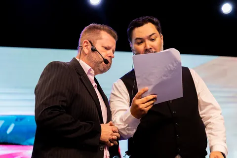 Clayton Yaso repassando o roteiro impresso com o co-apresentador no palco, os dois de microfone de cabeça.